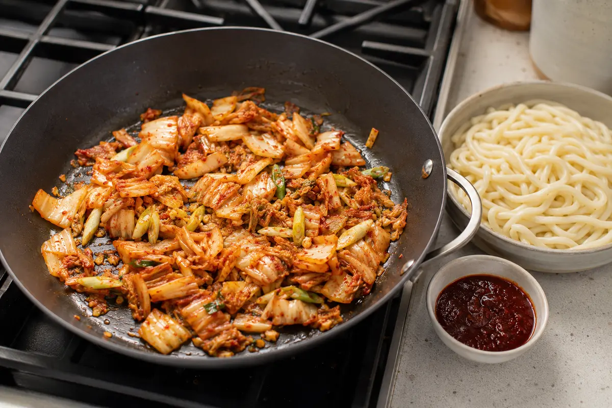 Kimchi and scallion whites cooking in a skillet beside a bowl of loosened udon noodles