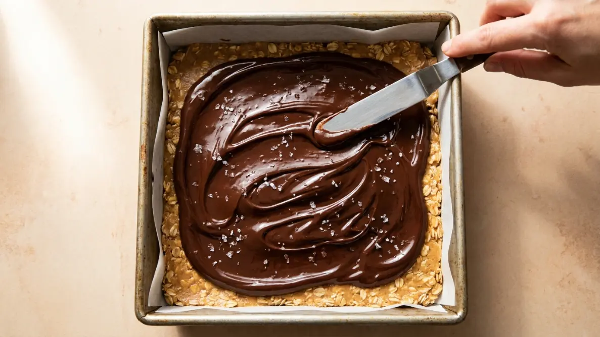 Melted chocolate being spread over the peanut butter oat base in a square pan