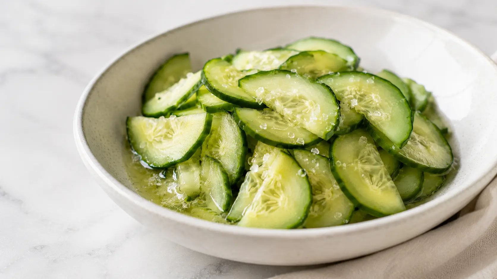 Thin half-moon slices of English cucumber salted in a white bowl before draining